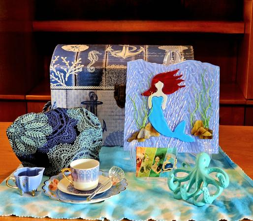 A tea tableau of an underwater scene of a mermaid by a sunken treasure chest, accompanied by a turquoise octopus, with waves & seaweed and a scallop shell for a teaspoon. The tea is a green tea flavored with apple pieces & flower petals.