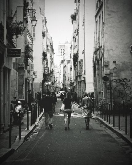 A black and white photograph of a narrow street in Paris. The street is lined with tall, old buildings adorned with balconies and street lamps. Three people are walking along the street, away from the camera. The street is bordered by bollards, and a parked scooter is visible on the left. In the distance, a larger building can be seen peeking above the rooftops.
