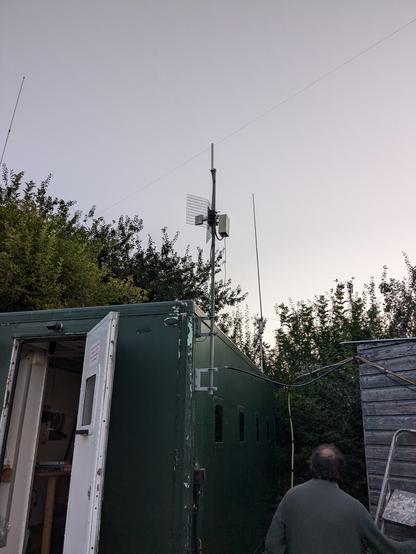 A green cabin with a pole mounted on the side in the foreground. 
On the pole: a 2.4ghz directional antenna (WiFi) and a very small colinear (ADSB).
In the background, an X300 colinear is visible.
There are some cables on the right hand side, hanging down by the side of the cabin, in the process of being run towards the cabin.