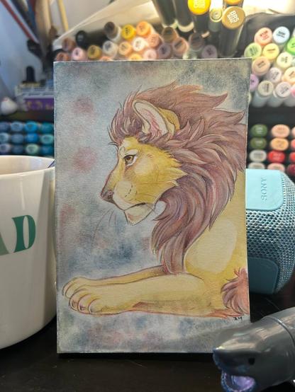 A picture of a lion painted in watercolor. He's looking left and is seen in a profile view. His fluffy main is a darker red brown shade while he is a more tan shade of brown.