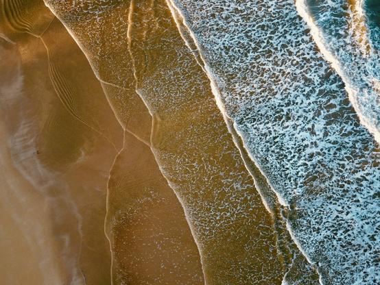Fotografía aérea a color de una playa, donde las olas del mar rompen suavemente sobre la arena dorada, creando un contraste entre los tonos azulados del agua y los tonos cálidos de la tierra.
