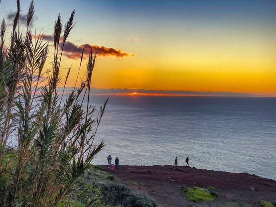 Das Bild zeigt die im Meer untergehende Sonne mit einem blau zu gelb-orange verlaufenden Himmel mit einzelnen Wolken oben links im Bild. Im mittleren Vordergrund schaut man auf einen abfallenden, felsigen Hang, auf dem zwei Pärchen stehen, bzw. gehen. Am linken Bildrand, ganz im Vordergrund, stehen ein paar Büschel Schilf- oder Pampasgras.