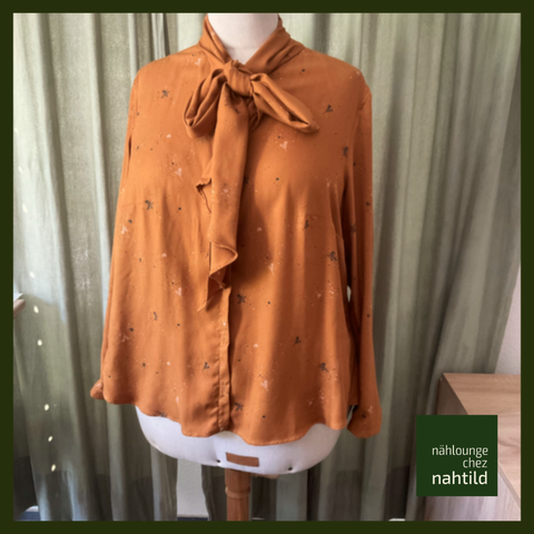 Siennafarbene Bluse mit kleinen grünen Elementen auf einer Schneiderbüste. Die Bluse hat eine Schleife am Halsausschnitt vorne in der Mitte. Unten rechts ein Logo "Nählounge chez Nahtild". Im Hintergrund ein Vorhang.