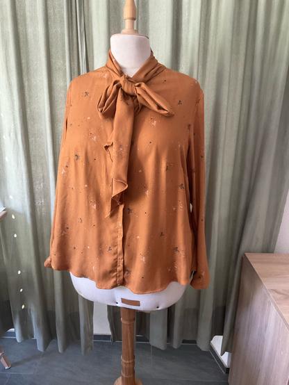 Siennafarbene Bluse mit kleinen grünen Elementen auf einer Schneiderbüste. Die Bluse hat eine Schleife am Halsausschnitt vorne in der Mitte. Im Hintergrund ein Vorhang.