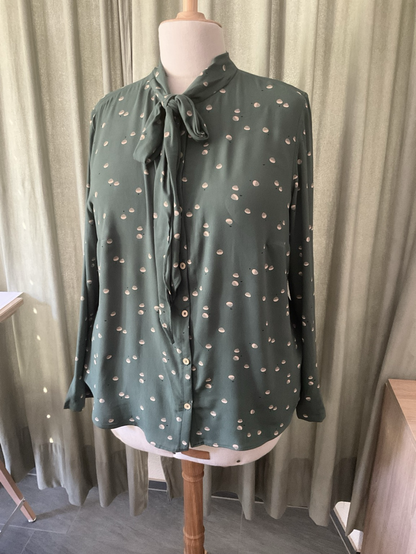 Grüne Bluse mit kleinen grünen Elementen auf einer Schneiderbüste. Die Bluse hat eine Schleife am Halsausschnitt vorne in der Mitte. Im Hintergrund ein Vorhang.