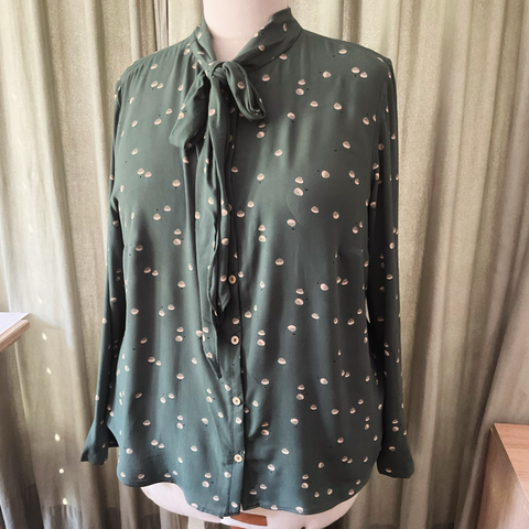 Siennafarbene Bluse mit kleinen beigen Elementen auf einer Schneiderbüste. Die Bluse hat eine Schleife am Halsausschnitt vorne in der Mitte. Im Hintergrund ein Vorhang.