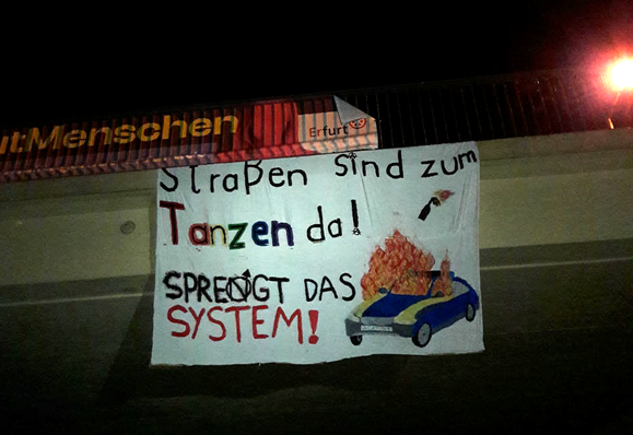 Ein Banner hängt von einer Brücke, darauf steht "Straßen sind zum tanzen da! Sprengt das System!" mit einem gemalten, brennenden Auto.
