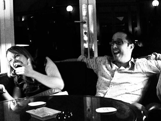 A black and white photograph captures a candid moment between two individuals seated on a sofa. The woman on the left is laughing heartily, her hand partially covering her face in the midst of her laughter. The man on the right is also laughing, his arm draped over the back of the sofa. The background is dimly lit, but a glass table in front of them reflects some of the ambient light.