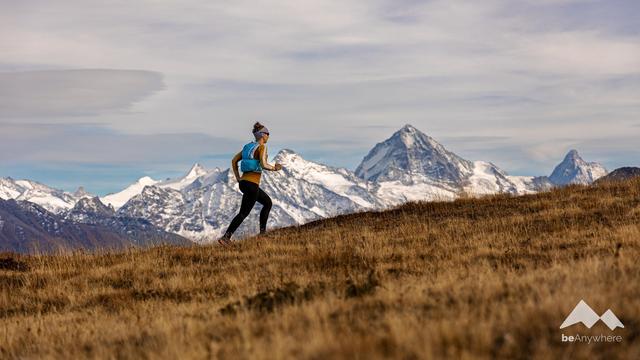 Women wearing running clothes and a running backpack, she's running upwards with huge alpine mountains in the background. Peaks are covered with snow.
