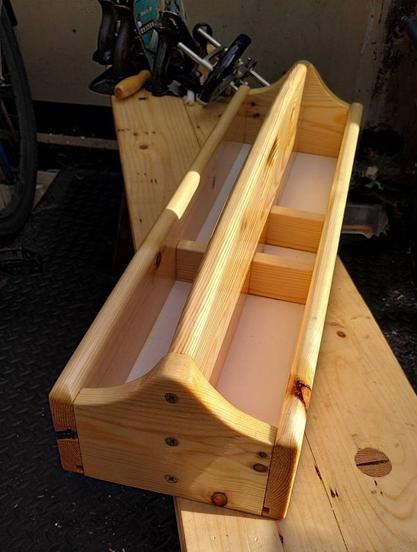 A wooden tool box, long and narrow with a handle down the middle. One side has two storage areas and the other has three. The box sits on a wooden bench with hand planes visible in the background.