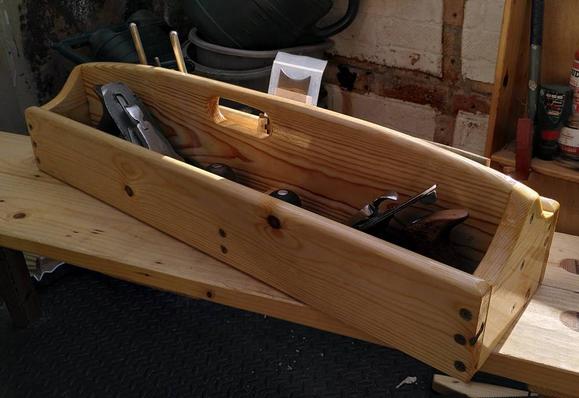A wooden tool box, long and narrow with a high handle down the middle. Two hand planes can be seen in the near side, and other tools are just visible on the far side. The box sits on a wooden bench with clutter visible in the background.