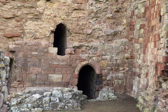 A stone wall, constructed from squared blocks. The surface has come away on much of the wall, leaving an irregular surface.
At the base of the wall, a small arched doorway leads down into the dark. Above and to one side, is a narrow, arched window, also dark.