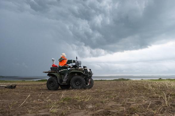 Eine Person in orangener Arbeitsregenjacke zieht mit einem Quad ein Metallrohr über einen Acker. Am Himmel sind Gewitterwolken zu sehen.