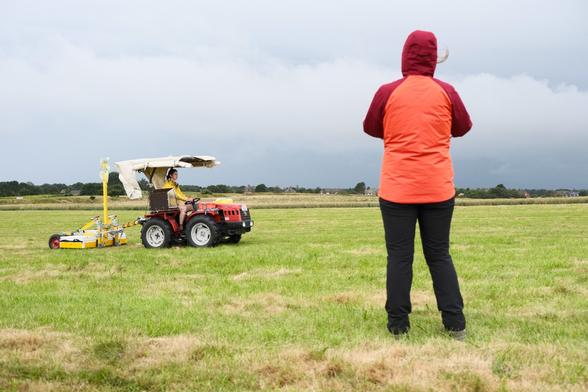 Ein kleiner Weinbautrecker zieht einen gelben Kasten über ein grünes Feld. Eine Person in roter Regenjacke steht im Vordergrund.