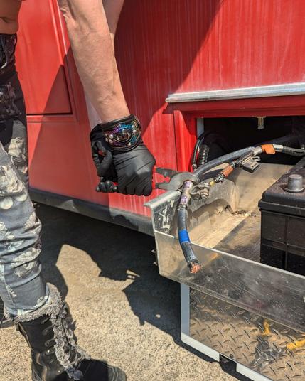 A person leans their bare arms and one leg into the photo, gripping wire clippers clamped around a hefty cable tightly. They are wearing black latex gloves and an interesting leather wrist cuff decorated with a painted glass eyeball. The cable runs next to a large automotive battery in a compartment coming off of a large vehicle.
