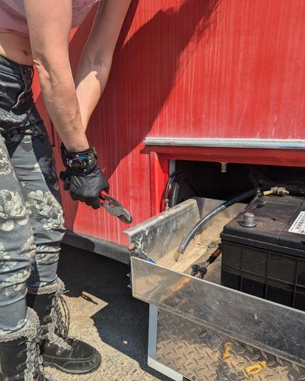 A person leans into the photo, showing only arms and legs, gripping empty wire clippers tightly. They are wearing black latex gloves and an interesting leather wrist cuff decorated with a painted glass eyeball. A clipped cable runs next to a large automotive battery in a compartment coming off of a large vehicle.