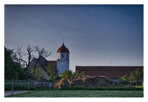 Im Vordergrund ein Teil einer Koppel, links die Einzäunung und Anschnitt eines Gebäudes, daneben ein abgestorbener Baum, die Dorfkirche und das Dach eines Gebäudes, dessen Mauern von einem grossen Misthaufen verdeckt werden.