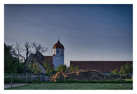 Im Vordergrund ein Teil einer Koppel, links die Einzäunung und Anschnitt eines Gebäudes, daneben ein abgestorbener Baum, die Dorfkirche und das Dach eines Gebäudes, dessen Mauern von einem grossen Misthaufen verdeckt werden.
