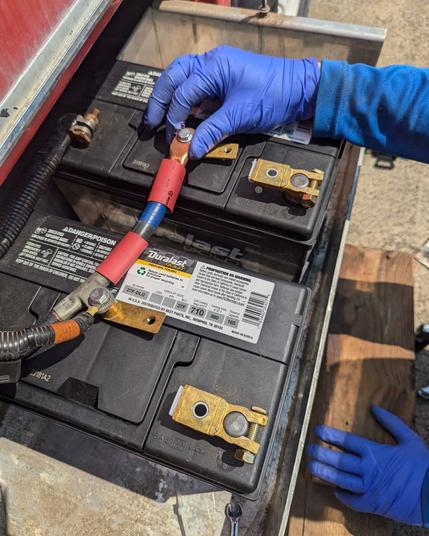 Two large automotive batteries with custom connectors attached are underneath a hand with blue gloves on holding a hefty battery cable sporting the other half of the custom connector.