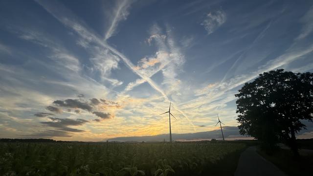 Sonnenuntergang mit einem Himmel voller leichter Wolken, Windkraftanlagen am Horizont und einem Feld mit Bäumen im Vordergrund.