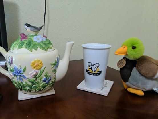 Foto de tekruĉo apud taso apud pluŝanaso. La taso havas emblemon de la programara projekto GNU. La anaso ĉarmas, kaj supozeble en sia kapo antaŭĝuas la teon! La flava tekruĉo estas el fajenco, kun multaj floroj k folioj malplatige metitaj sur sia flanko. Sur la fermilo estas elstara kvazaŭskulpta birdo sur lito el folioj kaj floretoj.