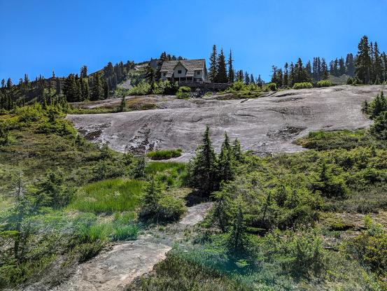 A lone small building stands atop a knob of glacially smoothed columnar andesite. Alpine evergreen trees mantle the skyline. The building is a fine example of CCC work. It's Region 6 rustic style includes: irregular plan, roof and porch projections, intersecting gables, dormers, multi-paned windows, and variations in exterior texture.