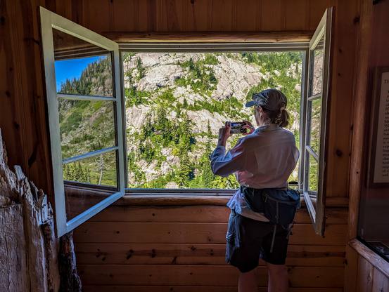 A woman gazes out an open window to take a photograph of mountain scenery.