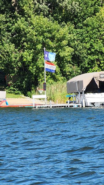 Flags spotted along a lake shore. Top one is new Minnesota flag. Bottom flag says Cold Beer. On the right, a pontoon boat is docked.