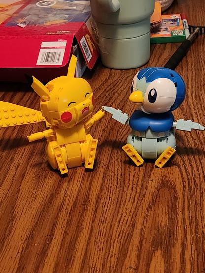 Photo. Building block recreations of Pikachu and Piplup, posed to appear as though they are seated and holding hands. Pikachu, left, is smiling with its eyes closed.