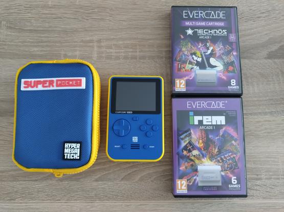 Photo de la console portable Super pocket.
A gauche de la console, une sacoche de rangement de couleur bleu et jaune. En haut, un bandeau blanc ou il est écrit "Super pocket" en rouge.
En bas à droite, le nom du constructeur "Hyper Mega Tech!" en blanc sur un fond noir
A droite deux boîtes de jeux. Ce sont des compilations des jeux d'arcade de IREM et Technos. Les boîtes représentent divers artworks des jeux présents dans les cartouches entourées par un cadre violet. En haut, il est écrit "Evercade".
Au milieu, la console portable "Super Pocket édition Capcom" de couleur bleu sur la face avant et jaune sur les côtés et le dos.