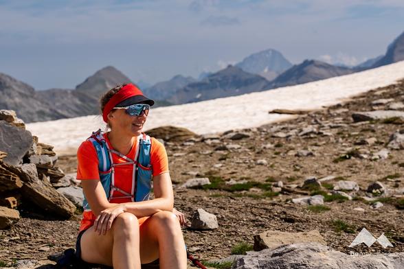 Woman wearing her trailrunning gear sitting between boulders in the Swiss mountains and taking a break from her trail run. Snow and high peaks in the background.