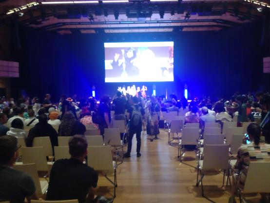 Blick in den Hauptsaal, der sich nach Ende des Cosplay Wettbewerbs grade leert. Im Hintergrund auf der Bühne posieren die Teilnehmerinnen und Teilnehmer für ein Gruppenfoto.