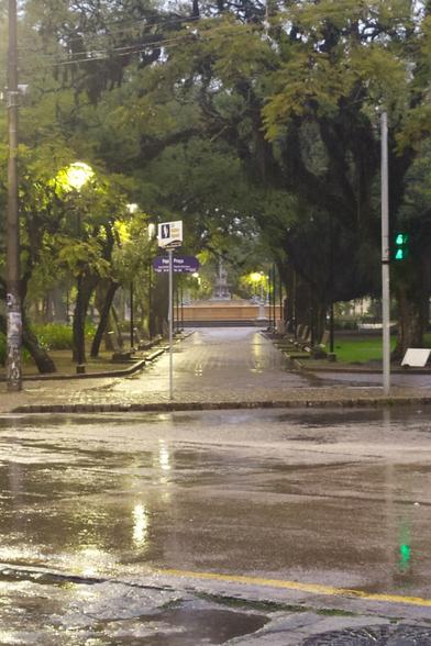 Foto de uma das entradas para a praça Coronel Pedro Osório, caminho de paralelepípedos, com bancos ao longo do caminho, nas laterais, até a parte central, onde tem um chafariz. Caminho coberto por árvores, que ficam logo atrás dos bancos. Tempo chuvoso,