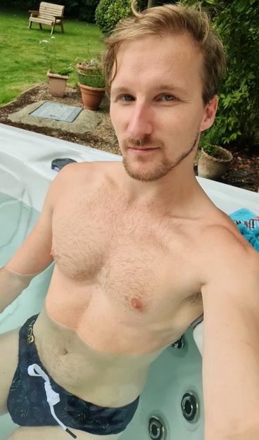Image of a blonde haired male sitting in a hot tub with dark green patterned swimwear on. A garden with a bench is visible in the background.