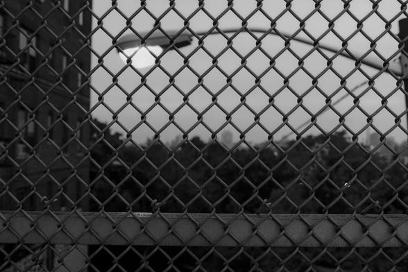 A black and white photo of a fence by Riverview Park in The Heights, Jersey City. Behind the fence is a streetlight, then the trees at the top of the heights, and a hazy and foggy outline of the NYC skyline in the distance.