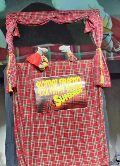 A puppet stage draped in red and white tartan fabric, with a sign on the front saying SCOTTISH FALSETTO SOCK PUPPET THEATRE SUPERHEROES in yellow font.  In the opening are two puppets.  the one on the left is dressed as Thor, and the one on the right is wrapped in the label from a can of IRN BRU
