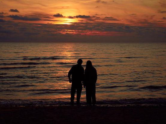 Silhouette of two individuals standing on a beach at sunset, with the sun low on the horizon casting a warm glow and reflecting off the water.