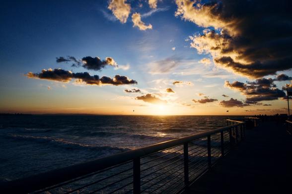 Ein Sonnenuntergang. Mit Wolken. Bild ist in orange blau gehalten. Am Himmel sind Möwen zu sehen und ein Drachenflieger. Der Steg und die Ostsee sind sehr dunkel bis schwarz gehalten um eine Silhouette darstellen zu können.
