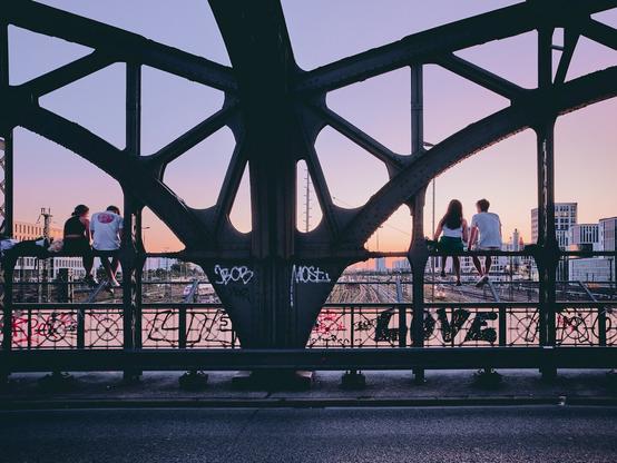 Das Bild zeigt eine Szene auf der Hackerbrücke in München während des Sonnenuntergangs. Vier junge Menschen sitzen auf dem Stahlgerüst der Brücke und blicken auf die darunterliegenden Bahngleise. Die Silhouetten der Personen heben sich gegen den farbenfrohen Himmel ab, der in warmen Tönen von rosa bis lila leuchtet. Die Brücke ist mit Graffiti bedeckt, und im Hintergrund sind moderne Gebäude und Stadtstrukturen zu erkennen. Die Atmosphäre wirkt ruhig und friedlich, während die Menschen den Ausblick genießen.