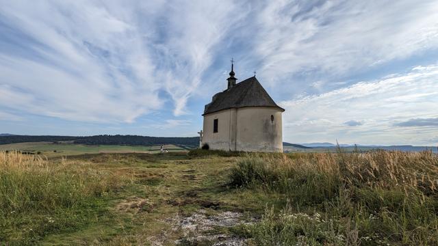 A small chapel, white round with dark grey roof and one small tower on the far side, on flat hilltop, yellow green grass, lime stone spots, other flat hills in the background, white clouds on the blue sky in the shape of radiant stripes emitted from the chapel.