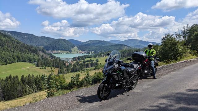 Two motorcycles, one with rider, standing on the side of the road above a distant lake worth turquoise color and forest covered hills in the background. Blue sky, scattered entre clouds.