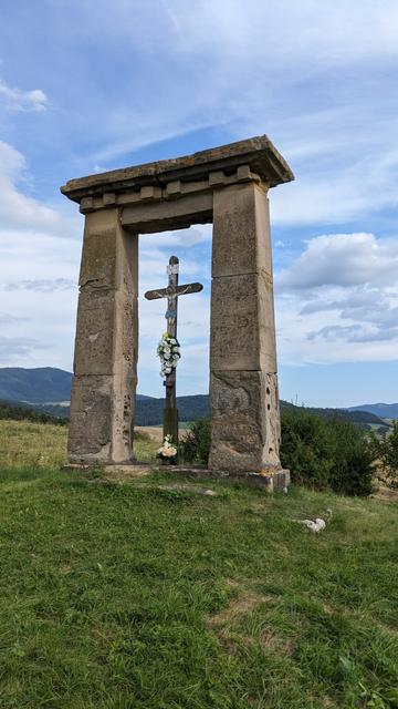 Llime stone rectangular arch with wooden cross in the space inside, green grass, green bush behind one pillar, forest covered hills in the background.