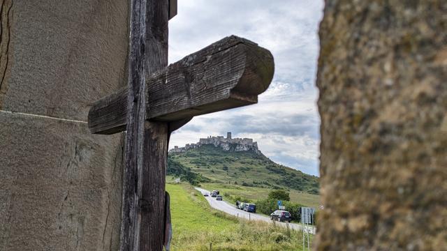 View through the before mentioned arch on the other side, pillars on the side of the photo, part of the cross with arms. Under one arm in the distance is a castle on the hill with a road approaching it. Green grass around, sky covered with white clouds.