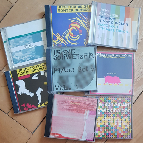 Eight CDs by Irene Schweizer spread out on wooden floor "Chicago Piano Solo", "Irène Schweizer/Günter Sommer", "To Whom It May Concern", "Irene Schweizer/Louis Moholo", "Piano Solo Vol. 1", "John Tchicai-Irène Schweizer Group: Willi the Pig", "Ulrichsberg" with Pierre Favre, and "Spring" with Jürg Wickihalder