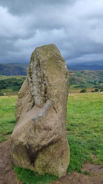 A two metre high monolith stone, with grassland and pretty fells in the distance. The grass is dotted with yellow flowers, and the sky is grey.