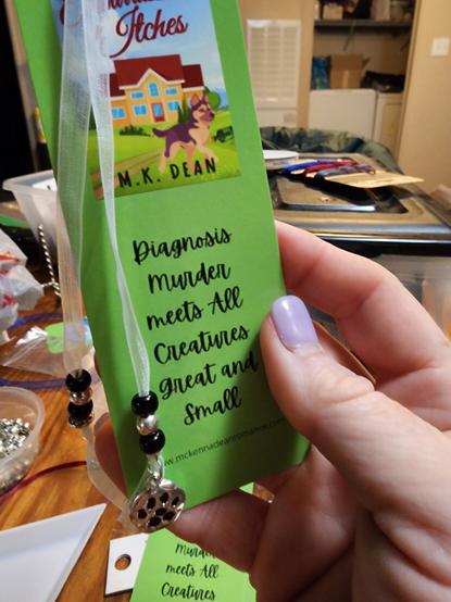Grass green bookmark with the cover of An Embarrassment of Itches by M.K. Dean: Diagnosis Murder meets All Creatures Great and Small, white ribbon, alternating black and silver beads, with a pawprint charm