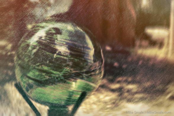 An experimental image of a green gazing ball before a shed. The entire image is faded and worn.