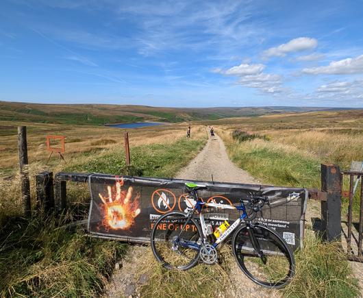 A bicycle in front of a metal gate with a gravel road behind the gate. The gate has a large poster covering it and a large, fiery hand is visible to the left of the poster. The location is moorland and the sky is blue and cloudless.