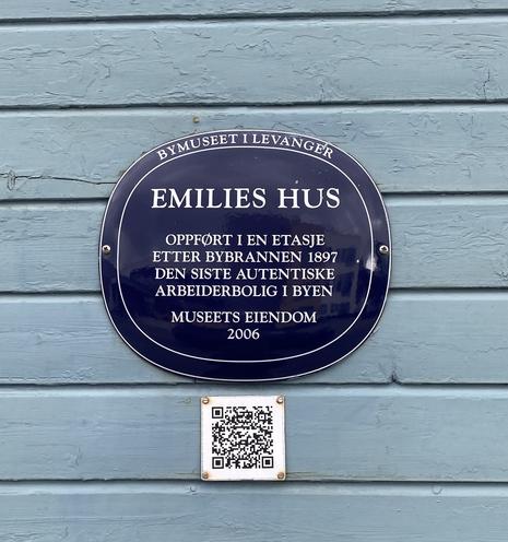 Et blått avrundet informasjonsskilt om huset, med en QR-kode under. På skiltet står: EMILIES HUS. Oppført i en etasje etter bybrannen 1897. Den siste autentiske arbeiderboligen i byen. Museets eiendom 2006.