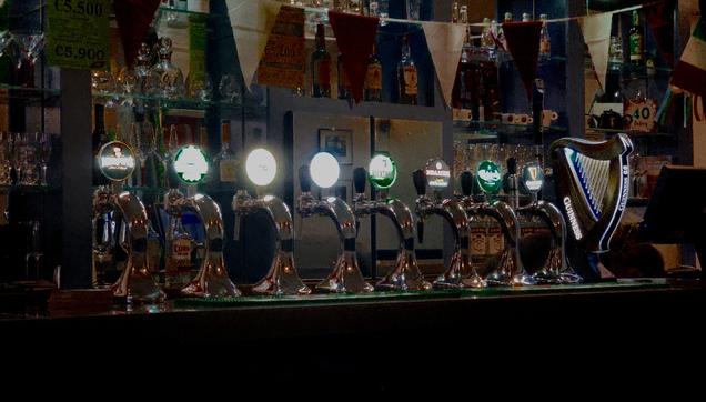 Barra de pub irlandés. 8 tiradores de cerveza en fila con el logo redondo de cada marca arriba, aunque en algunos no se distingue por los reflejos. A la derecha de la fila hay una figura del logo de Guinness, con el arpa irlandesa. Salvo los reflejos, la escena tiene poca luz.